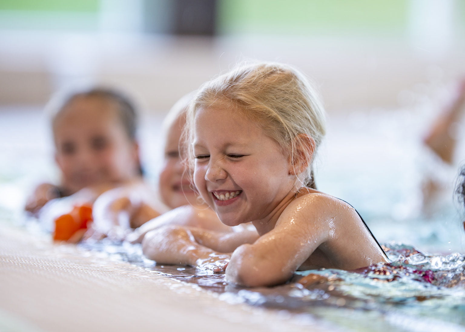 A group of girls are attending a swimming class. One of them is holding onto the side of the pool and laughing while kicking.